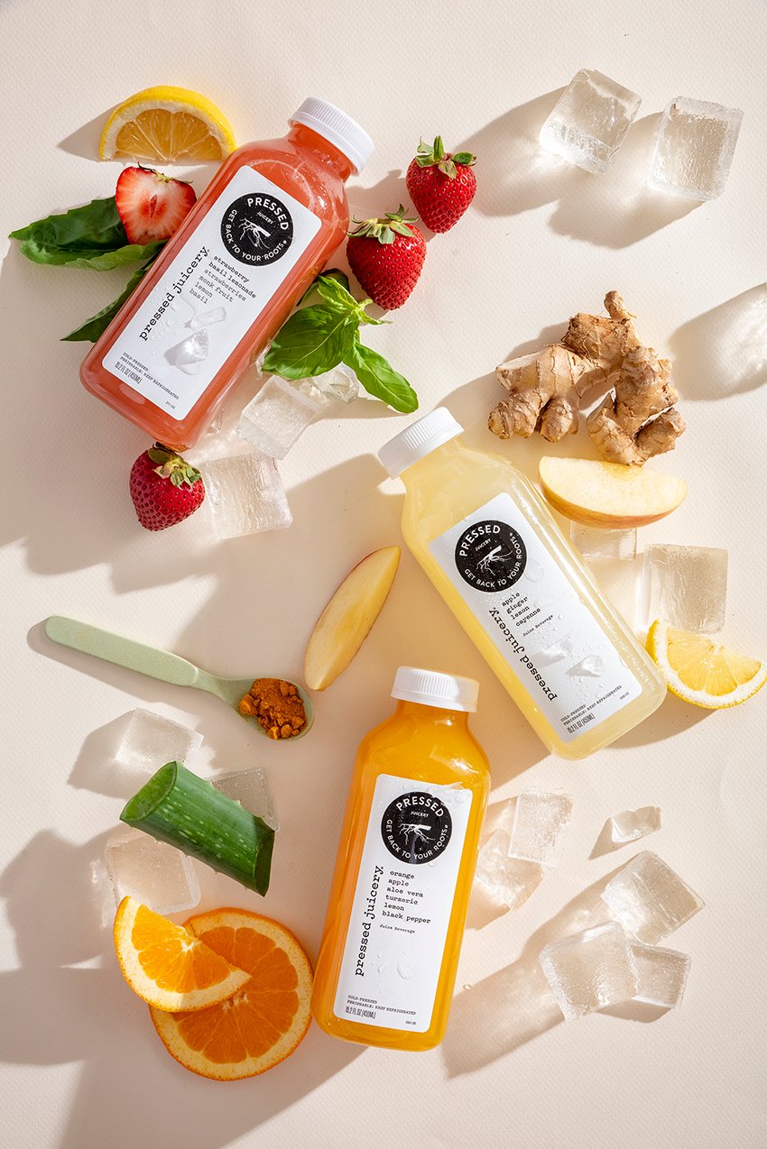 Two bottles of juice from the Pressed Juicery displayed with a piece of aloe vera, ice cubes, orange slices, lemon wedge, apple slice, spoon full of turmeric powder