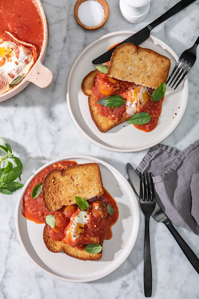 A plate of poached eggs on toast smothered in tomato sauce and garnished with fresh basil and black pepper, accompanied on the table by a sprig of basil, black fork and knife, and a grey cloth napkin