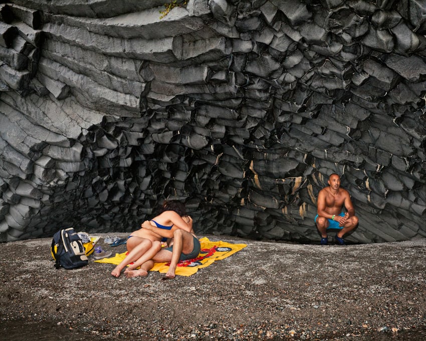 A single man sits under a rock formation behind a couple kissing. 