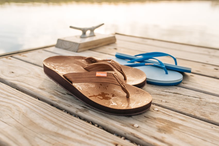 Photographer Jillian Clark's photo for FeelGoodz featuring two pairs of flip flop sandals on a dock with calm water in the background. One pair is brown and is wet, and the other pair, behind those, is blue.