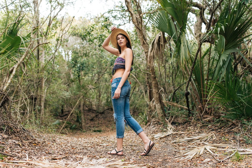 Photographer Jillian Clark's image of a woman wearing cropped denim pants, a crop top, a brimmed straw hat, and strappy black sandals looking over her left shoulder. She is standing in a tropical forest. 