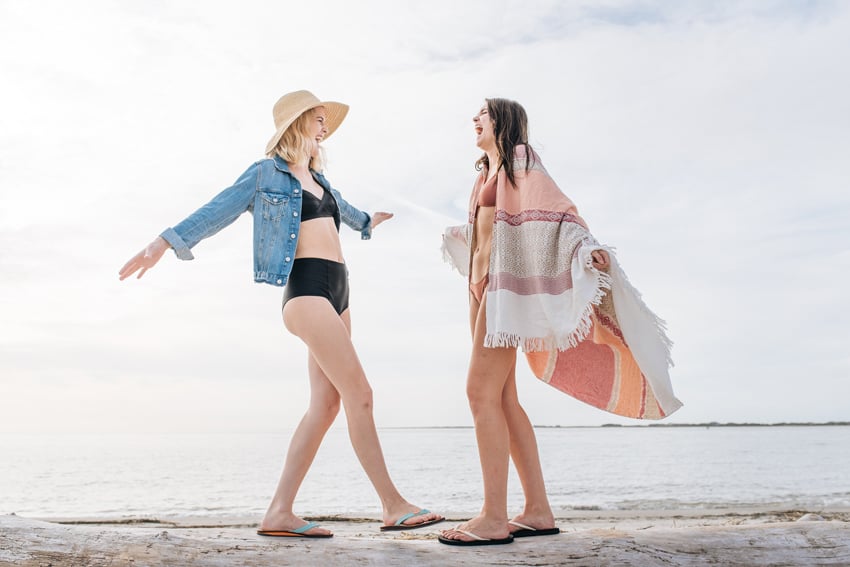 Jillian Clark's photo for FeelGoodz featuring two women in bathing suits and flip flops laughing together. They are on a beach and are both standing on a log with the ocean in the background.