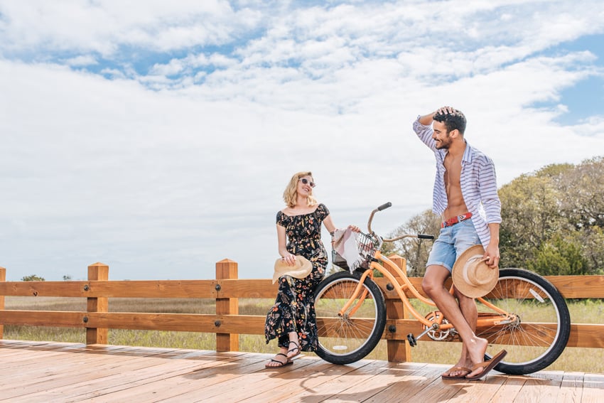 Jillian Clark's photo for FeelGoodz featuring two people towards the right of the frame on a boardwalk with a bicycle smiling at each other. The woman wears a long black dress with a colorful print and strappy black sandals, and the man wears denim shorts and an open button-down shirt with leather thong sandals. Each holds a sun hat in one hand.