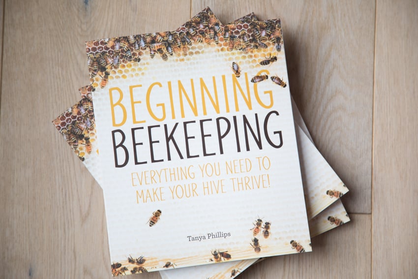 A photo of a stack of three copies of Tanya Phillips' book, "Beginning Beekeeping - Everything You Need to Make Your Hive Thrive!" The cover of the book features a honeycomb covered with bees.