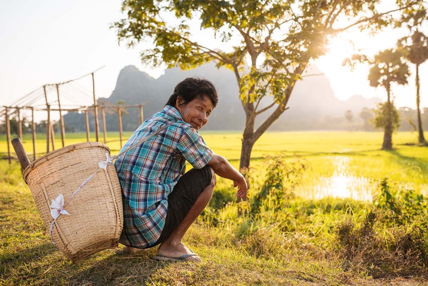 A photograph by Ben Pipe of a Burmese man squatting in grass with a large basket strapped to his back. He looks over his right shoulder, smiling gently.