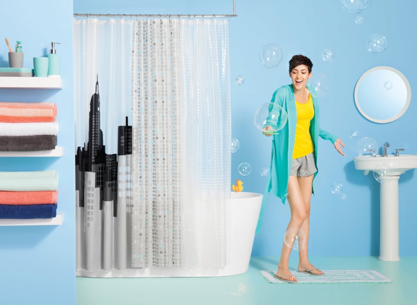 A photo for Target by Paul Owen of a woman in a staged bathroom set. She stands next to a bathtub with a shower curtain around it that has a city skyline design on it. She is surrounded by large bubbles and is catching the largest one on a bubble-blowing wand in her right hand. She has a pixie haircut and wears a bright yellow top, gray shorts, a short teal bathrobe, and gray flipflop sandals.
