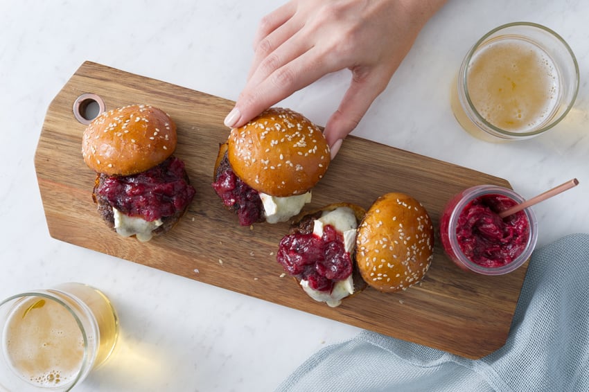 Morgan Ione's photo for Bon Appétit Magazine featuring Président brand brie. The photo features three sliders with golden, sesame seeded buns on a wooden serving board. The buns are each slightly off-kilter, revealing burger patties with melted brie and a deep pink condiment on top. A person's hand is in the shot touching the slider in the center. A container of the pink condiment sits on the right. On the upper right and lower left are glasses of golden beer.