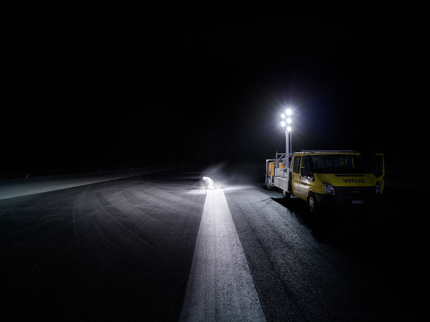 A photo taken by Scanderbeg Sauer of a person in protective clothing doing work in the night on an expanse of tarmac. The person works under a bright light from a fixture of a work truck parked nearby.