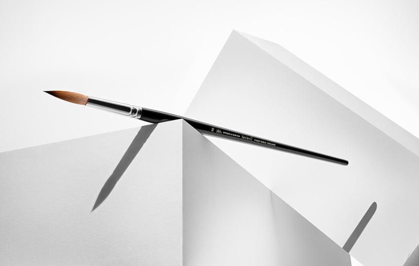 Timothy Hogan's photo for Winsor & Newton featuring a paintbrush with a black handle and metal ferrule. The paintbrush is balancing on the corner of a white box.