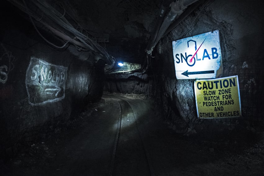 Luther Caverly's photo for Carleton University of an underground passageway to SNOLAB. The passageway is dark, with a single light visible and there are signs that say SNOLAB with arrows.