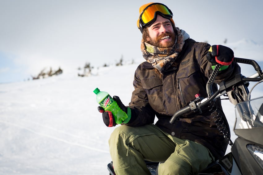 Photographer Adam Moran's photo for Mountain Dew. The photo features a man holding a green plastic bottle of Mountain Dew lounging off the side of a snow mobile in the snow. He is dressed for outdoor winter sports and wears his reflective goggles on his forehead.