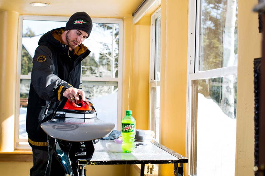 Photographer Adam Moran's photo for Mountain Dew. The photo features a man indoors in outdoor winter clothes. He appears to be polishing a snowboard, and there is a green plastic bottle of Mountain Dew on a table next to him.