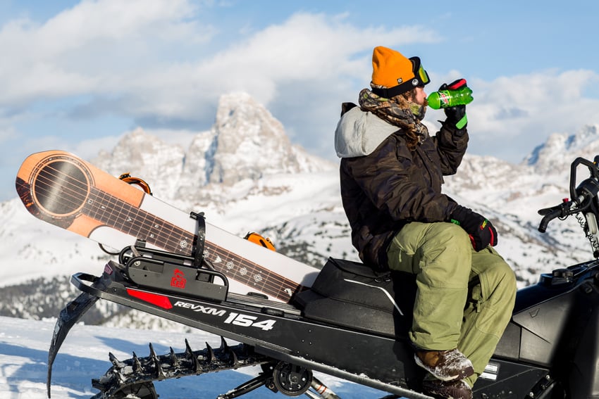 Photographer Adam Moran's photo for Mountain Dew. The photo features a man drinking from a green plastic bottle of Mountain Dew as he looks into the distance. There are snowy mountains in the background. He sits with both legs swung over one side of a snow mobile in the snow. A large snowboard that has a guitar design on it is strapped to the back of the vehicle.