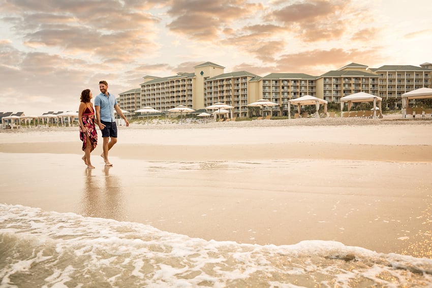 A romantic couple strolls hand in hand along the shoreline, complemented by the backdrop of a luxurious hotel complex. Photo by Amy Mikler.