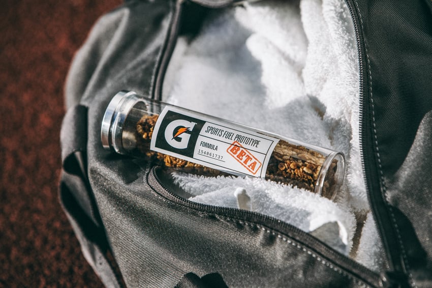 G L Askew II's image for Gatorade of their new granola snack product in a beaker style clear bottle package that sits on a towel inside an open sports equipment bag. The label says "Sports Fuel Prototype, Formula BETA."