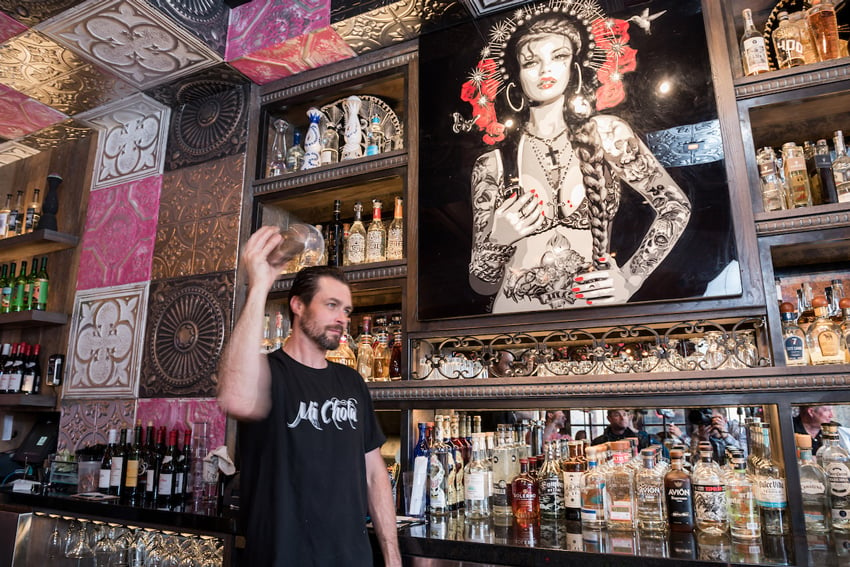A photo by C2 Photography for Aspen Sojourner Magazine. The image features a man at a bar shaking a cocktail shaker. There is an interesting piece of artwork behind him featuring an illustration of a heavily tattooed woman with a long braid and a halo. The wall and ceiling feature interesting large tiles.