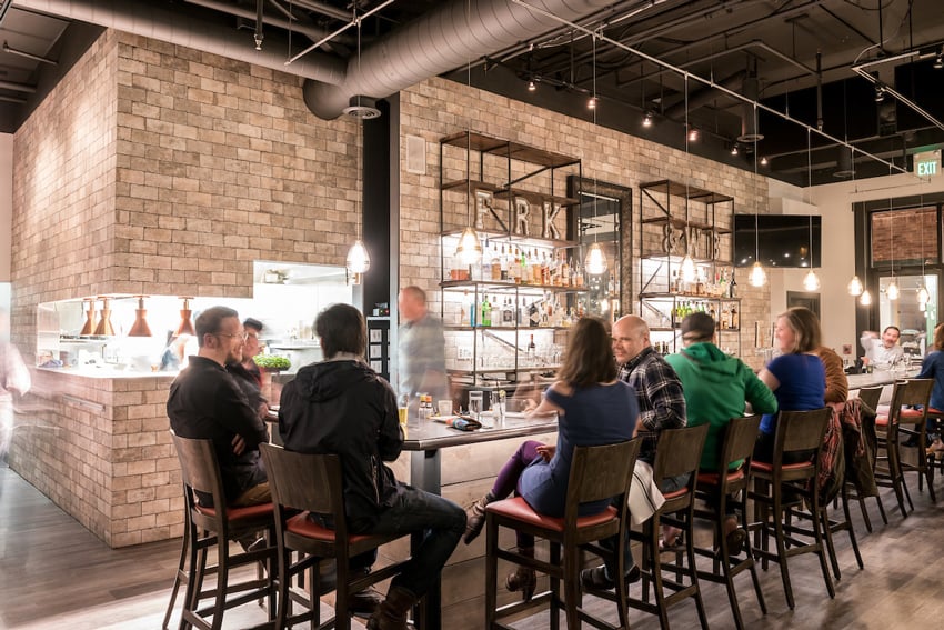 A photo by C2 Photography for Aspen Sojourner Magazine of a bar scene. Several people sit in tall seats at a modern looking bar. Anyone in motion is slightly blurred.
