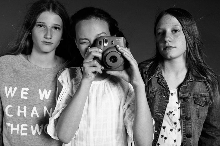 Photographer Caitie McCabe's black and white photo of three young girls taken in a studio setting. The girl in the middle squints into a polaroid camera aimed at the photographer, and the two girls flanking her look directly at the photographer with neutral expressions.