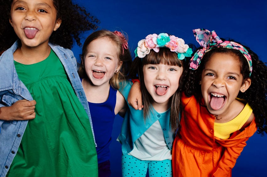 Photographer Caitie McCabe's photo taken in a studio with a cobalt blue backdrop. The image features four young girls wearing brightly colored clothing and characterful headbands. All four girls are sticking their tongues out playfully.