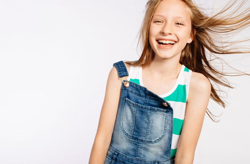 Photographer Caitie McCabe's image of a blonde-haired blue-eyed girl smiling. She is towards the right side of the frame and her straight hair swings spiritedly to the right. She wears a striped teal and white tank top under denim overalls with one strap unbuttoned. 