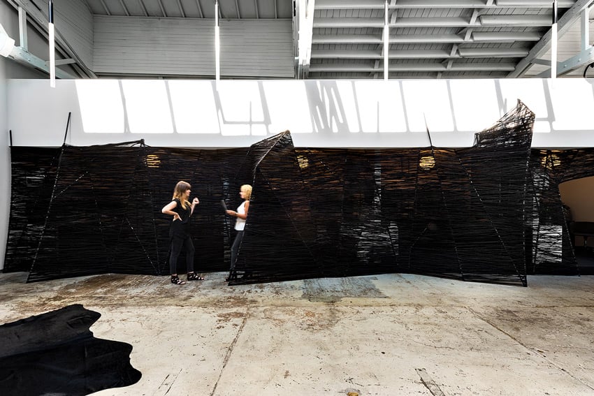 A photo by Chris Bradley for PROjECT Interiors of the interior of their office space. The photo features 2 woman standing in front of an interesting black screen that is partially transparent and almost looks like a black spider web stretched between poles that stand at various angles. Lighting fixtures from the space behind the screen show through. The floor is polished concrete.
