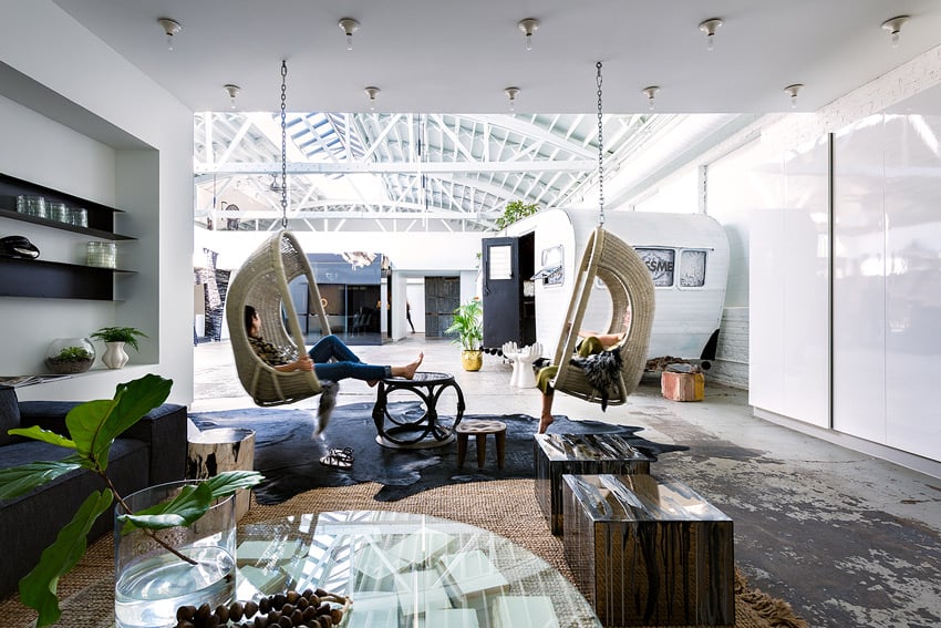 A photo by Chris Bradley for PROjECT Interiors of the interior of their office space. The photo features 2 barefooted women lounging across from each other in wicker swing chairs that hang from the ceiling. One woman rests her feet on a small wicker table in front of her. The walls are a bright white and there is a white trailer in the background.
