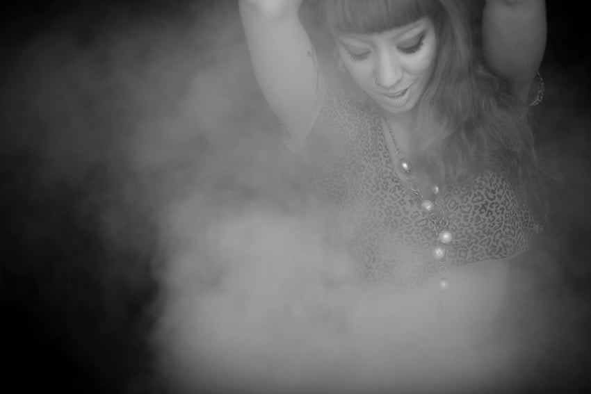 Claus Lehmann's photo featuring a woman with long wavy hair and bangs and heavy eye makeup dancing in a cloud of mist. The portrait is against a black background and the subject wears a short-sleeved cheetah print crop top and a long necklace with large pearls on it.