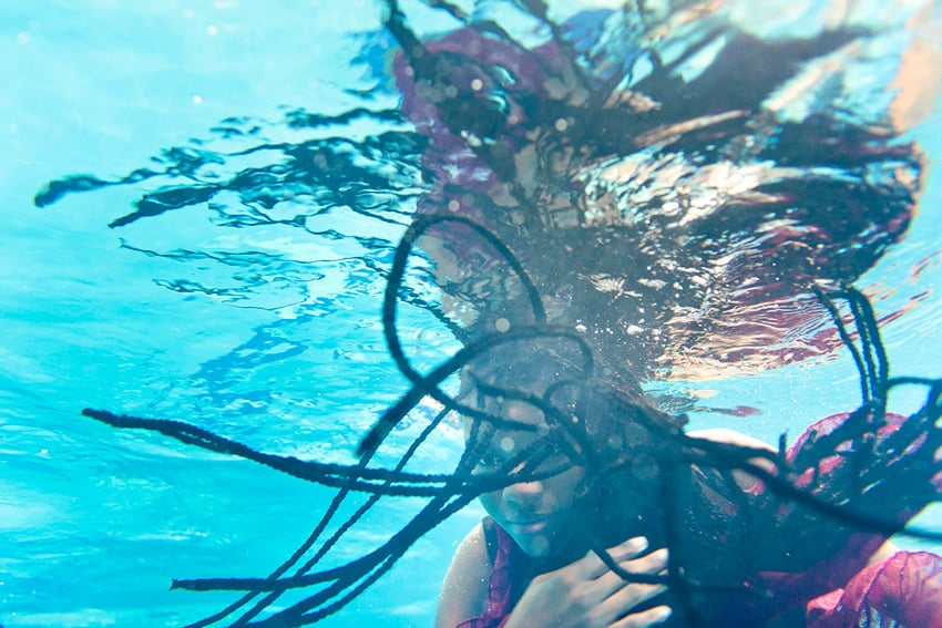 Claus Lehmann's portrait of a woman of color under water. She has long box braids that are suspended in the turquoise-colored water in front of her face. She wears a frilly fuchsia top.