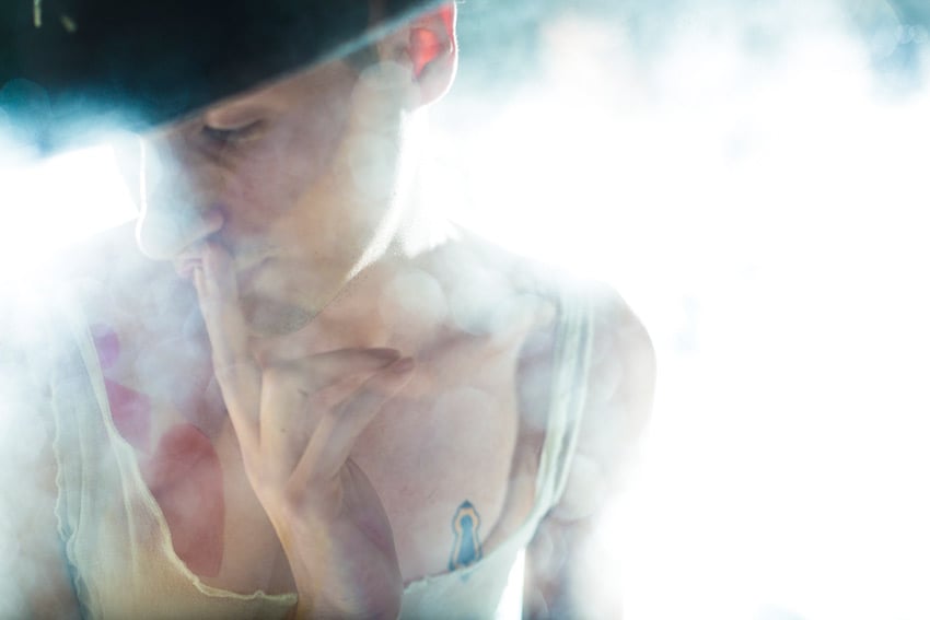 Claus Lehmann's portrait of a person with tattoos wearing a white tank top. The person also wears a black hat and is obscured by light and mist against a brightly lit background.