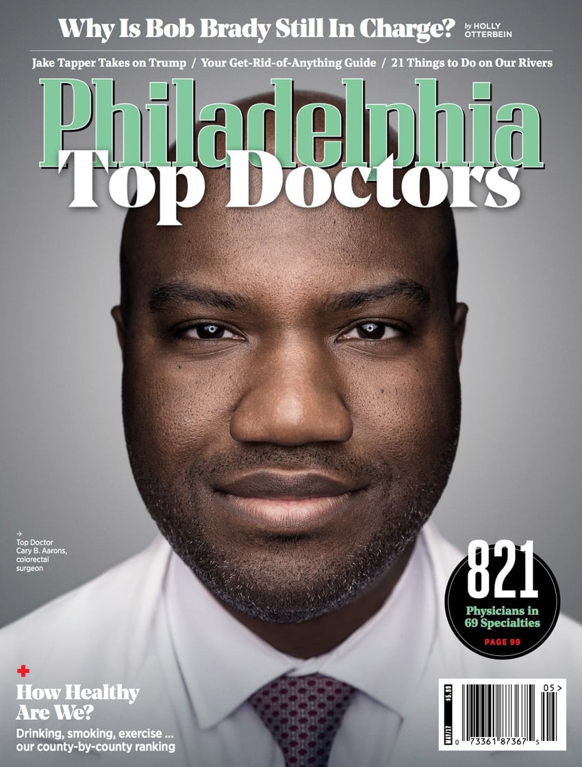 A headshot of Dr. Cary B. Aarons, a colorectal surgeon, by photographer Colin M. Lenton for the cover of Philadelphia Magazine. Cary is black and has short facial hair. He smiles without showing his teeth. Cary wears a white collared shirt with a maroon patterned tie under a white doctor's coat.