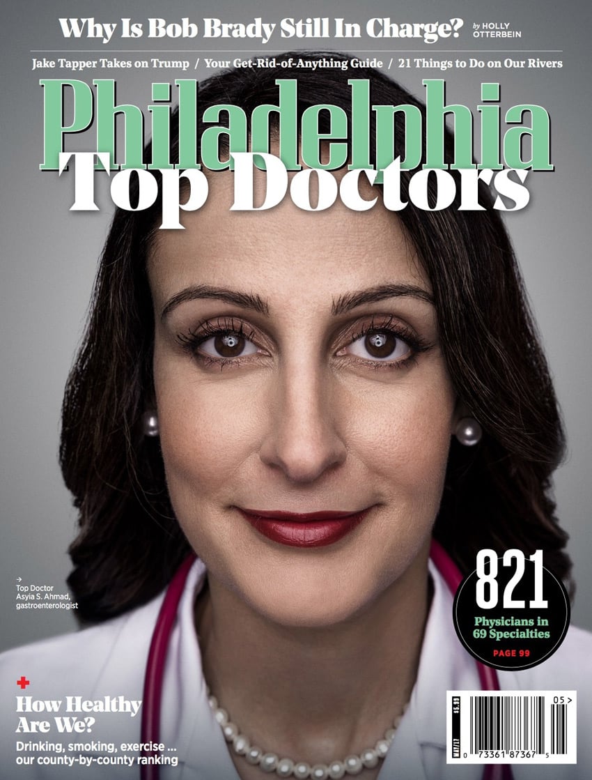 A headshot of Dr. Asiya S. Ahmad, a gastroenterologist, by photographer Colin M. Lenton for the cover of Philadelphia Magazine. Asiya is a woman of color. She wears pearl earrings, a pearl necklace, and a crimson lip color. She also wears a white doctor's coat and has a red stethoscope draped around her neck. Asiya smiles without showing her teeth.