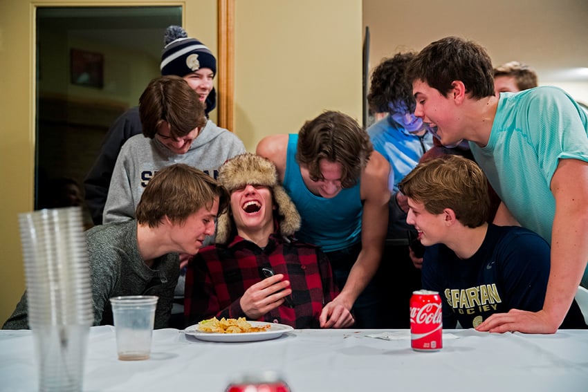 Photographer David Ellis' image of a group of boys laughing together at a table. The table in front of them has a can of Coca-Cola, a paper plate of potato chips, and a stack of clear plastic cups on it. 