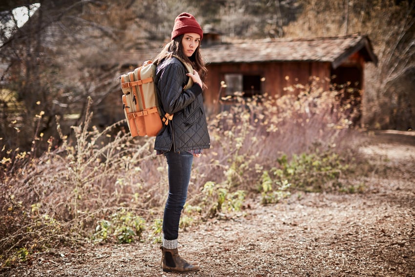 A photograph taken by David Troyer of a woman wearing an Eagle Creek backpack. She is wearing an outfit suitable for cold weather including thick socks, a beanie, and a quilted jacket, and she stands profile but looks directly at the camera.