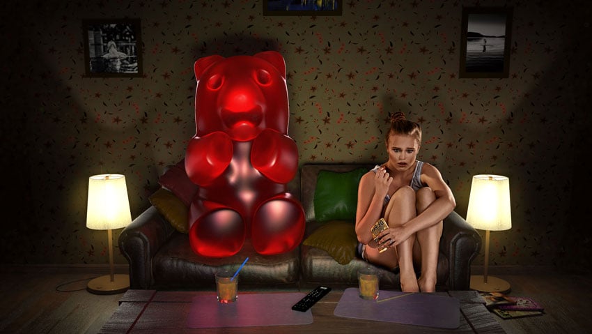 A photo by Diapolis Images for Haribo that features a woman with blonde hair sitting on a couch in a dark room with her knees to her chest holding a bag of Haribo gummy bears. Next to her on the couch is a life size red gummy bear. The two appear to be watching a movie together, and the woman has a frightened expression on her face.