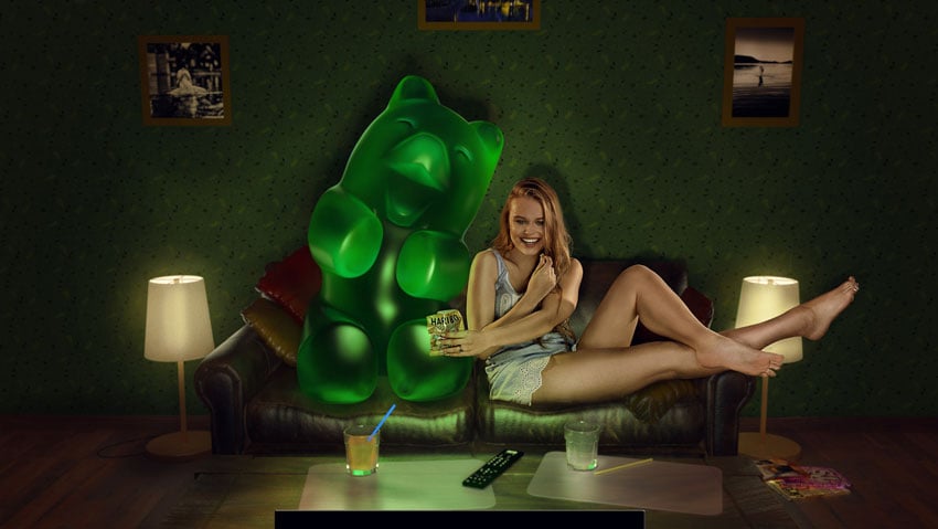 A photo by Diapolis Images for Haribo that features a woman with blonde hair lounging on a couch in a dark room. Next to her on the couch is a life size green gummy bear. The two appear to be watching a movie together, and the woman and gummy bear are laughing. She offers the bear a bag of Haribo gummy bears.