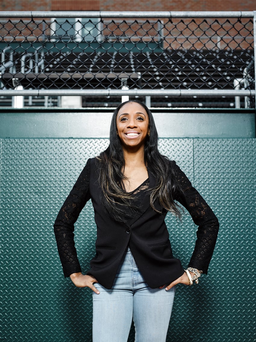 Doug Levy's portrait of Olympian Lashinda Demus for Financial Times. Lashinda wears her long hair down. She wears jeans and a black blazer and is posed, smiling warmly and looking off into the distance, with her hands on her hips in front of outdoor bleachers.