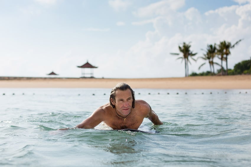 Ed Wray's photograph of paraplegic surfer Bruno Hansen paddling out on his surfboard . The water is a minty green and a sandy shore with palm trees can be seen in the background.