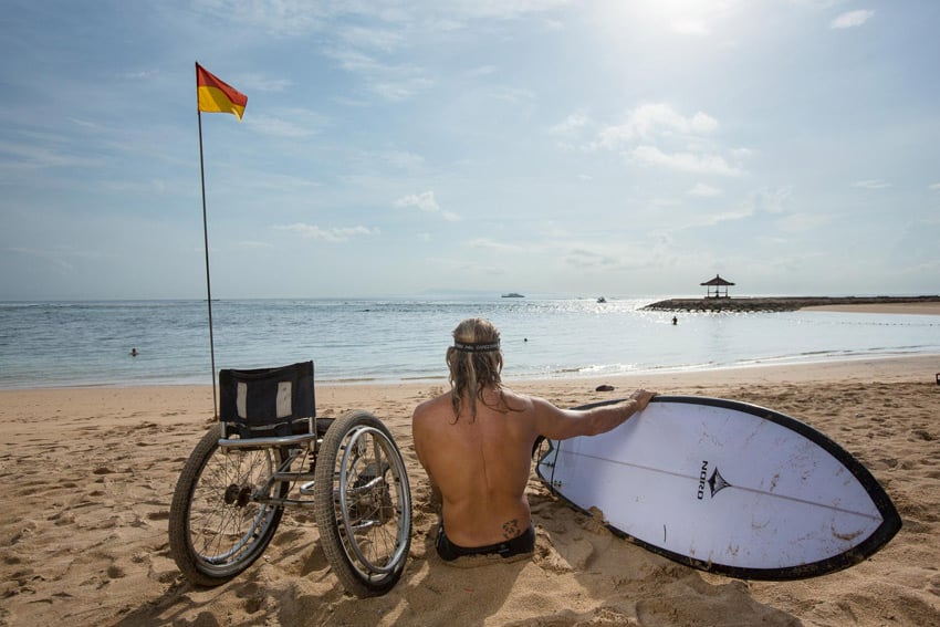 A photo by Ed Wray of Bruno Hansen from behind. Bruno sits in the sand on the beach in full sun between his white surfboard and his wheelchair. He looks out over the ocean. There is a flagpole in the sand to his left with a half yellow and half red flag.