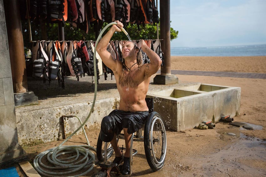 A photo by Ed Wray of Bruno Hansen in his wheelchair on the beach, rinsing his hair with water from a hose. There is a rack full of hanging life vests in the background.