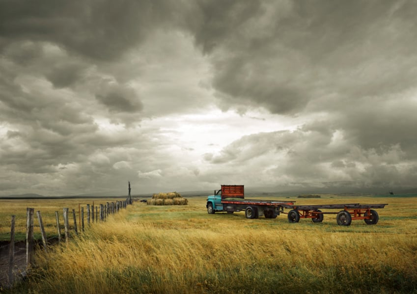 A grassy field with a working truck and hay bales under a moody sky, photo by Eric Schmidt