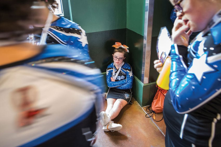 A young cheerleader sits in a corner of the room, anticipation evident in her posture as she waits, image by Felix Sanchez.