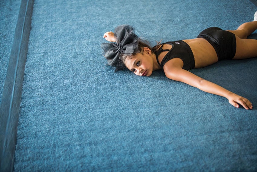 A young cheerleader lies on the floor, her form relaxed, perhaps reflecting on routines or preparing for the next performance with a sense of calm determination, photo by Felix Sanchez.