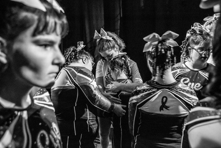 A black and white photo shows a group of cheerleaders backstage radiating solidarity as they hug each other, their smiles and embraces speaking volumes about their unwavering bond and teamwork, photo by Felix Sanchez.