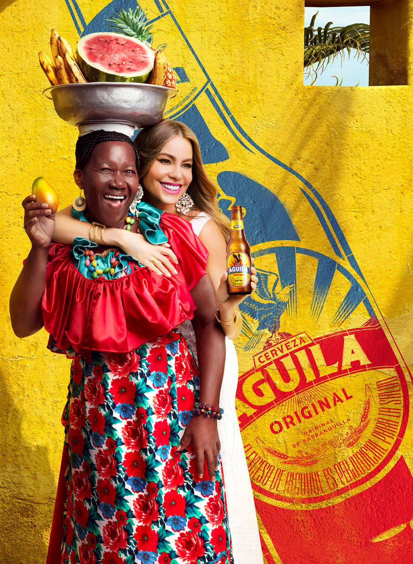 A photo by Fernando Decillis featuring Sofia Vergara embracing a woman from behind with one arm and holding a bottle of Aguila beer with the other. The woman wears a colorful dress and holds up a mango with one hand while she balances a bowl of tropical fruit atop her head.