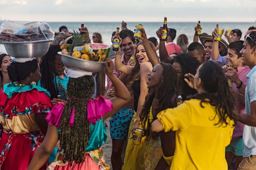 A photo by Fernando Decillis featuring Sofia Vergara toasting with a bottle of Aguila beer amidst a crowd of people in colorful clothing with the sea in the background.