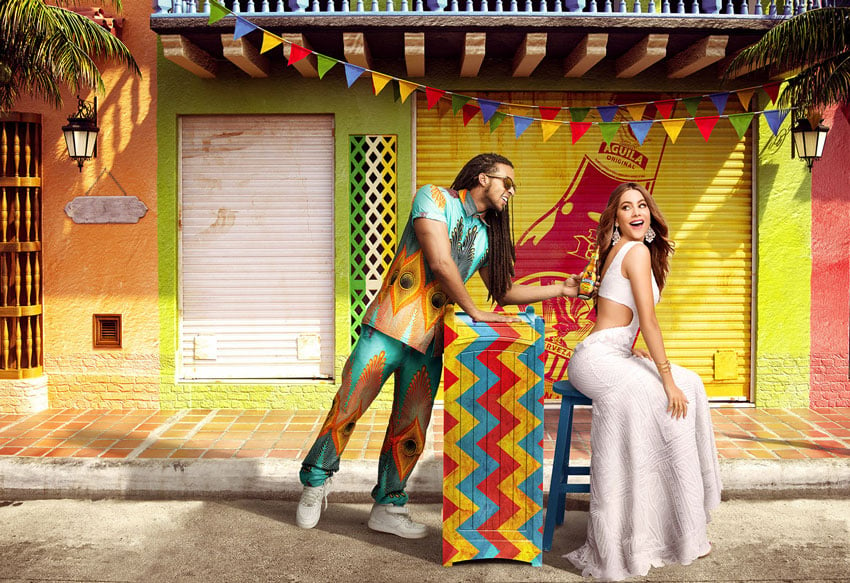 A color photo by Fernando Decillis of a man offering Sofia Vergara a bottle of Aguila beer in a tropical street setting.