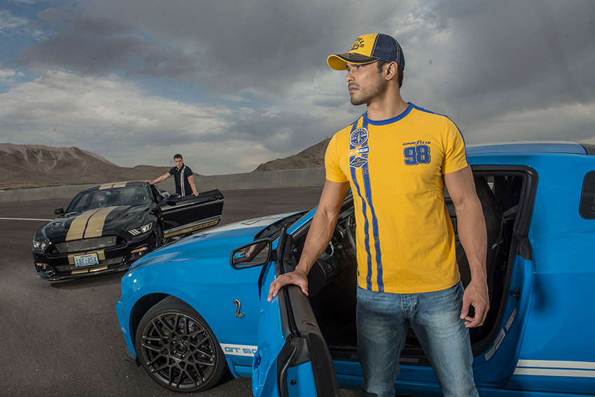 Photographer Jacob Kepler's photo for Goodyear Tire & Rubber Company.  The photo features a man in a yellow and blue Goodyear brand shirt modeled after a sports jersey and baseball cap. He stands in the open door of a blue sports car. In the background is a second man standing in the door of a black sports car with two broad taupe stripes down the center.