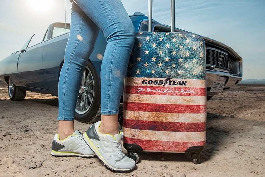 Photographer Jacob Kepler's photo for Goodyear Tire & Rubber Company.  The photo features a rolling suitcase covered in a fatigued American flag design and has the Goodyear logo across the center. Under the logo, it says "The Greatest Name in Rubber." Next to the suitcase is a person posing. Only their legs in slim-fitting jeans and feet in sneakers are visible in the shot. In the background is an old-fashioned blue convertible.