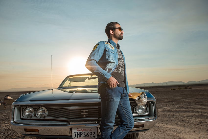 Photographer Jacob Kepler's photo for Goodyear Tire & Rubber Company.  The photo features a man leaning on the hood of an old-fashioned blue convertible with a California license plate. He models Goodyear brand clothing items and wears sunglasses. The sun sets behind him.