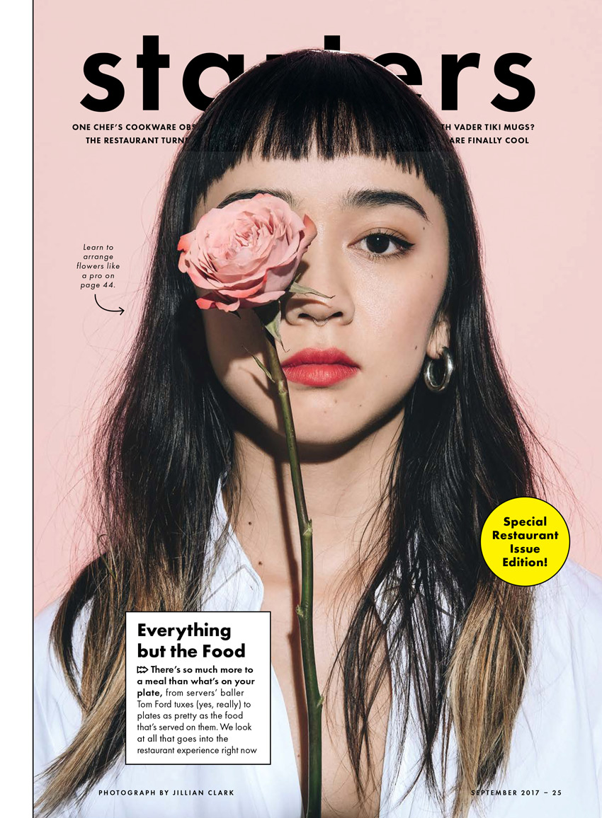 Jillian Clark's tear sheet for the cover of Bon Appétit featuring a woman with dark hair and bangs in a white shirt with pink lipstick against a light pink background. She looks directly ahead at the camera and holds a pink rose that covers her right eye.
